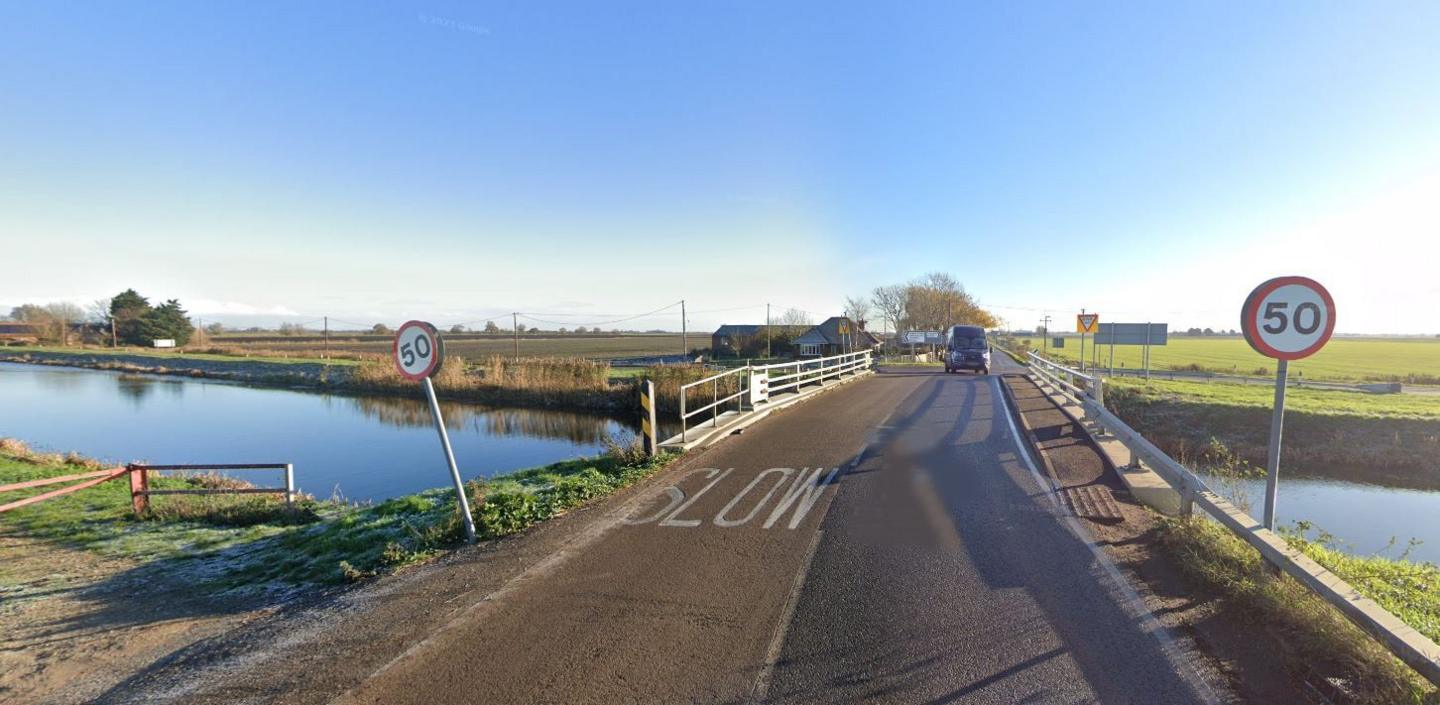 The Boots Bridge crossing the Sixteen Foot Drain, with a "slow" marking on the road surface, and a wonky 50mph sign on one side of the bridge