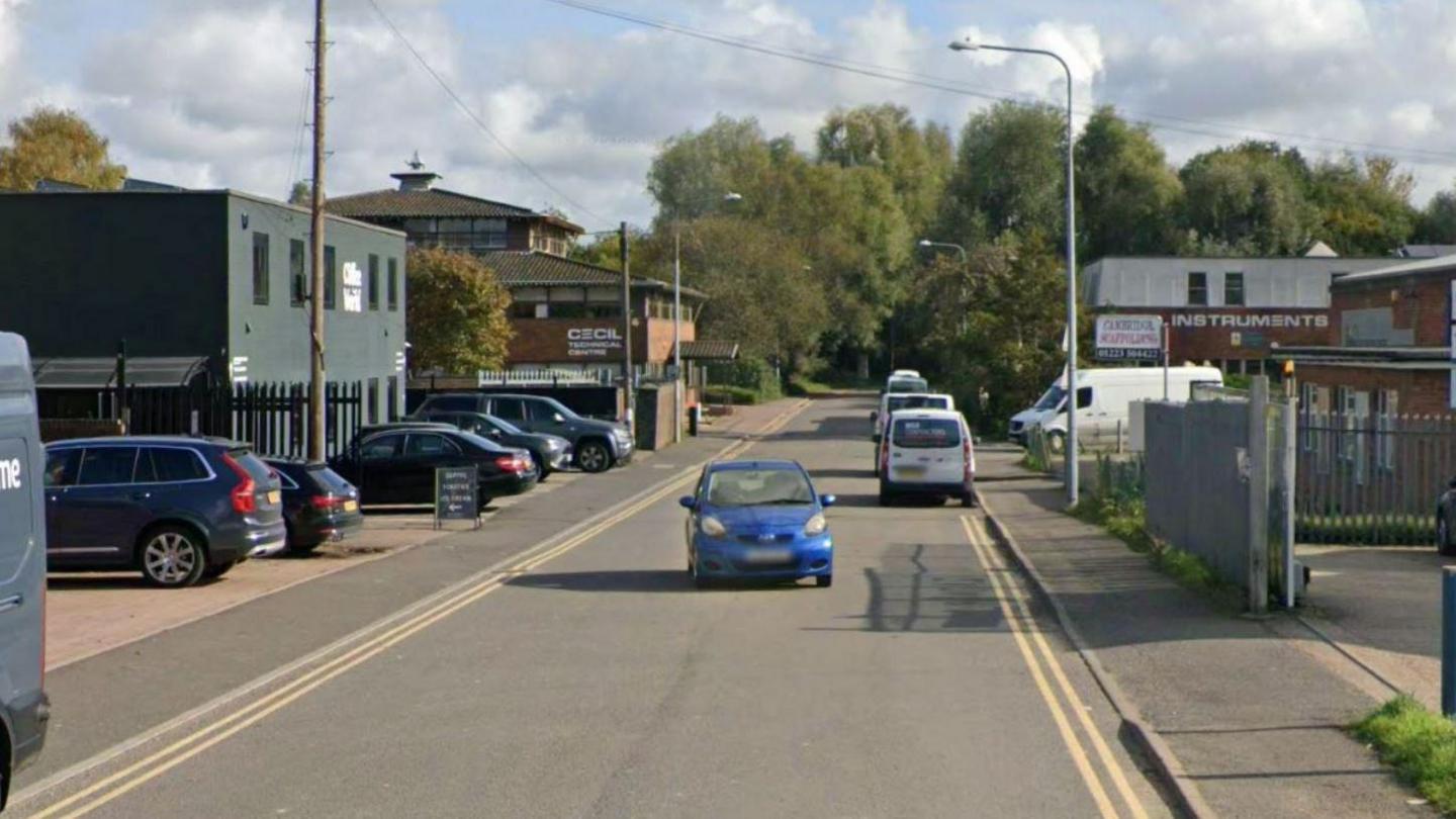 A road with double yellow lines on both sides. In the distance are some parked cars, and a blue car with its number plate blurred out is driving towards the camera.