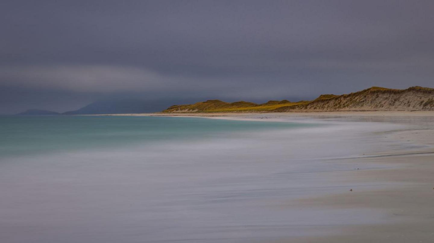 Green-blue sea, white sand and grassy sand dunes. In the distance are hills covered in low cloud.