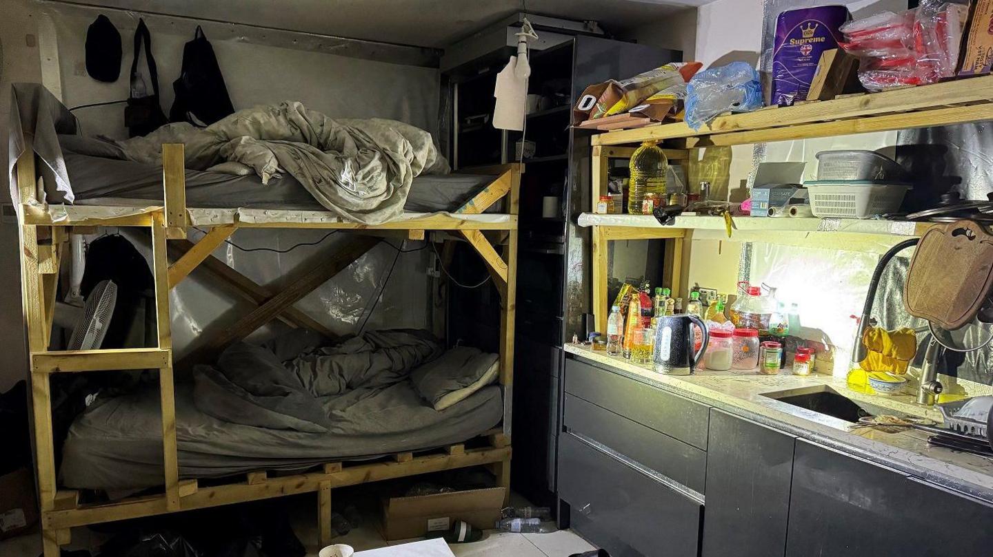 Bunk beds are in the corner of a kitchen area inside a house. They are scruffily-built and have untidy bedding on them. There are dark-coloured kitchen cabinets and a sink next to them. The worktops and shelves are stacked with food and plastic containers