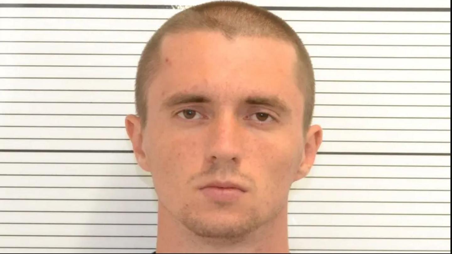 A mugshot of a man with a closely shaved head and brown stubble, with a serious expression. There is a white background with black horizontal lines.