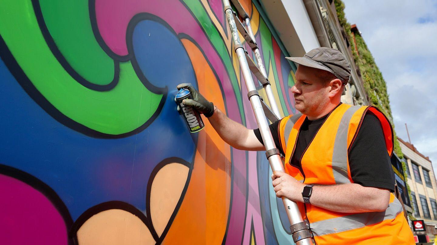 A man with a spray can in a high vis jacket spraying paint on a wall.