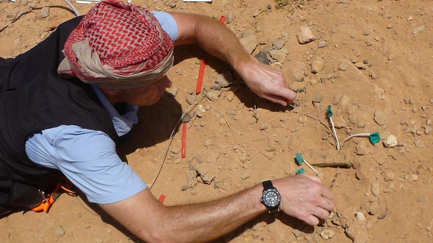 One of Gareth's pupils crawls on hands and knees towards a metal tin buried in the sand, with several white wires protruding. He is wearing a red and white headscarf.