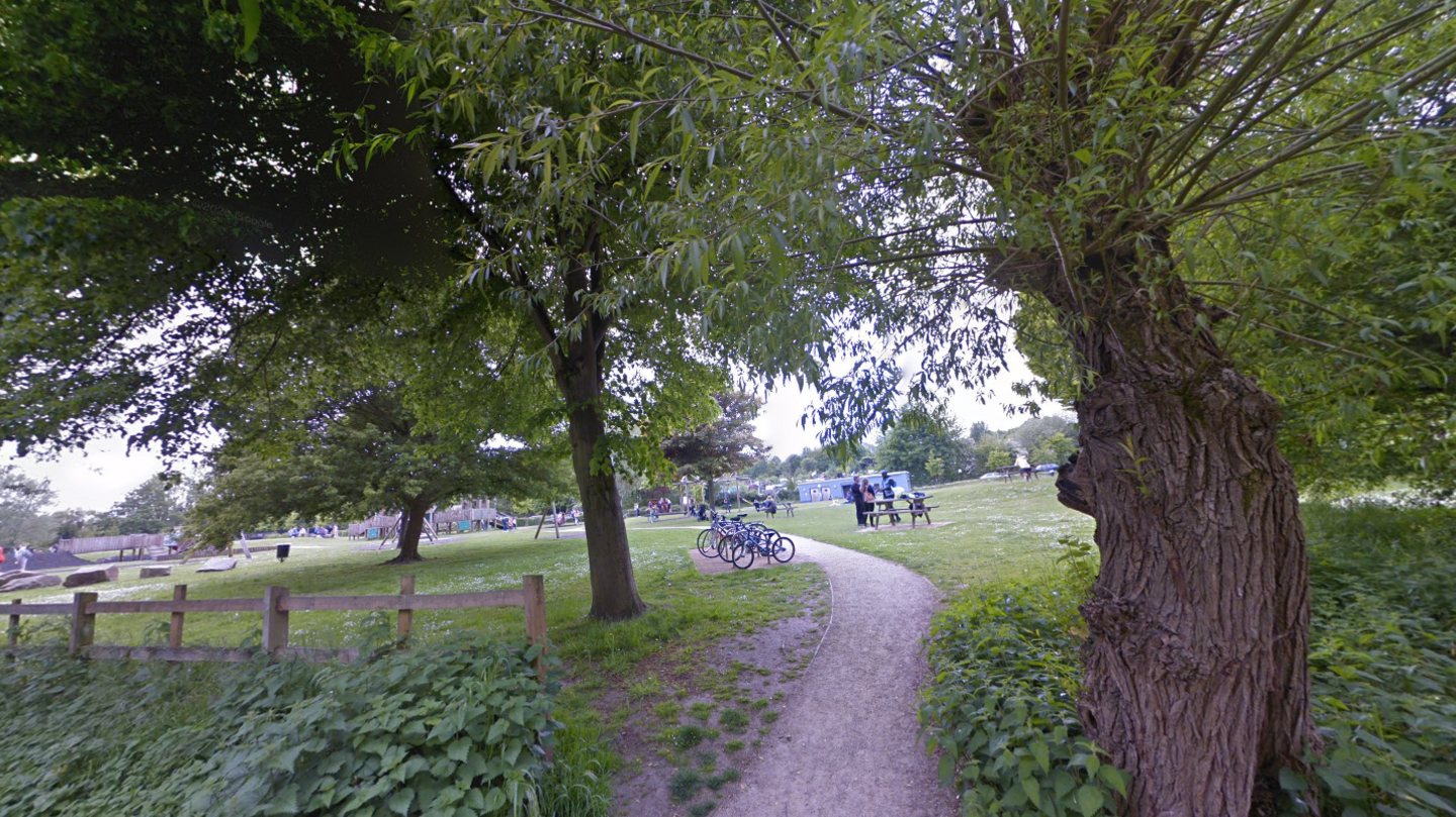 The entrance to Toddler's Cove playground in Whitehall Road, Canterbury, showing a path through trees, with bikes standing in the foreground and play equipment in the background.