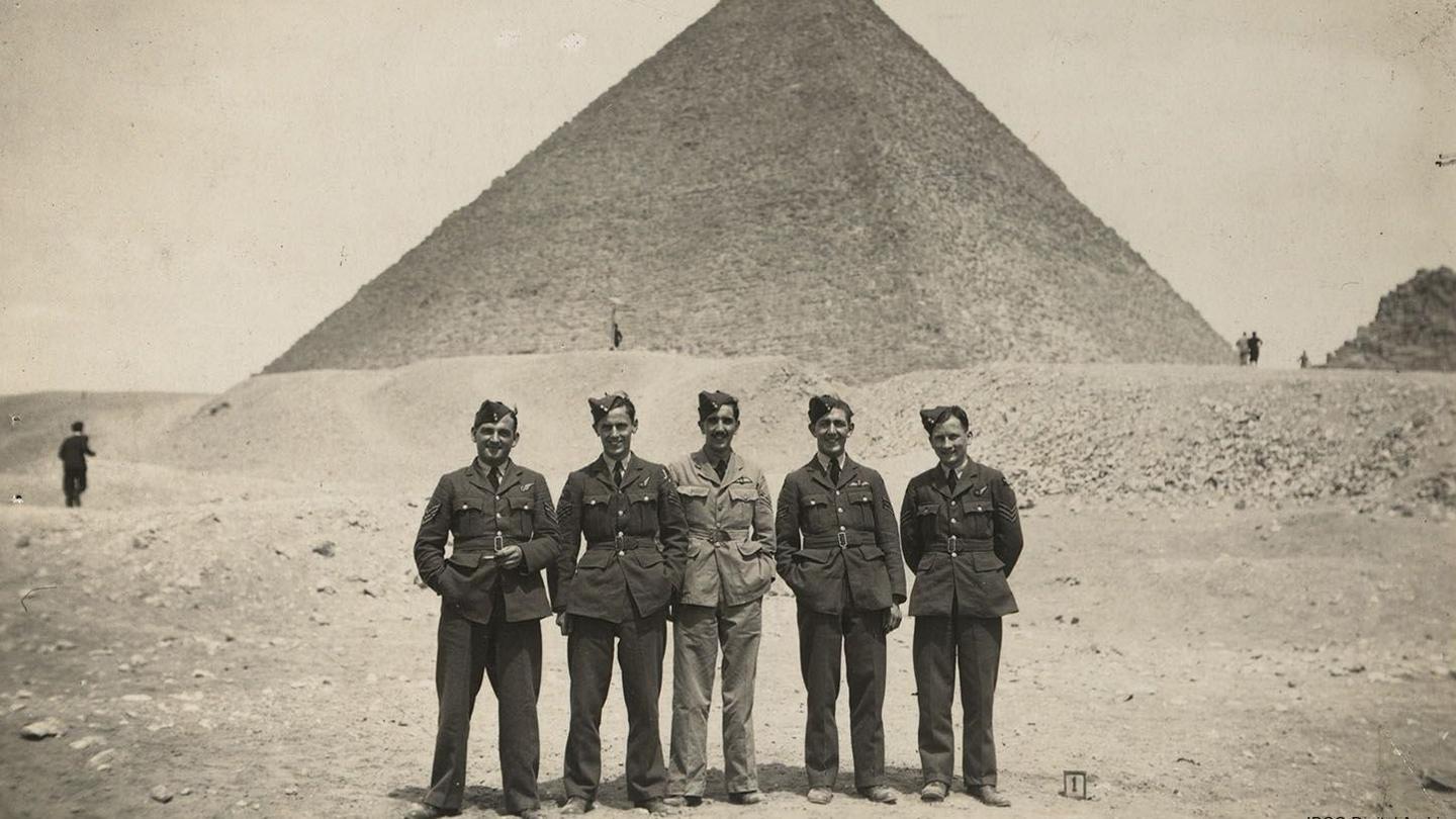 A group of five uniformed airmen, all wearing side caps, lined up in front of Great Pyramid of Giza (Pyramid of Khufu) at the Giza Necropolis, Egypt.