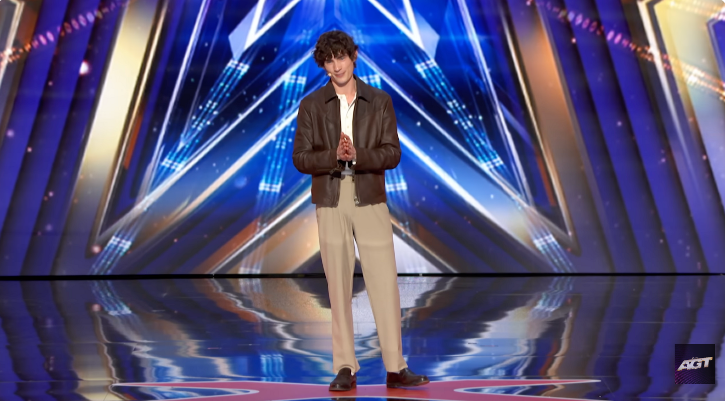 Joel is seen standing in the America's Got Talent stage, wearing a brown leather jacket and beige trousers. 