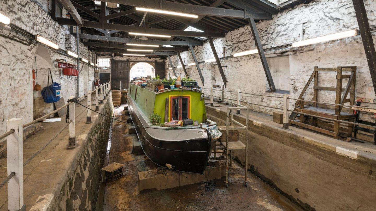 A green painted canal boat is propped up in a dry dock. There is strip lighting attached to the walls and ceiling joists of the building.