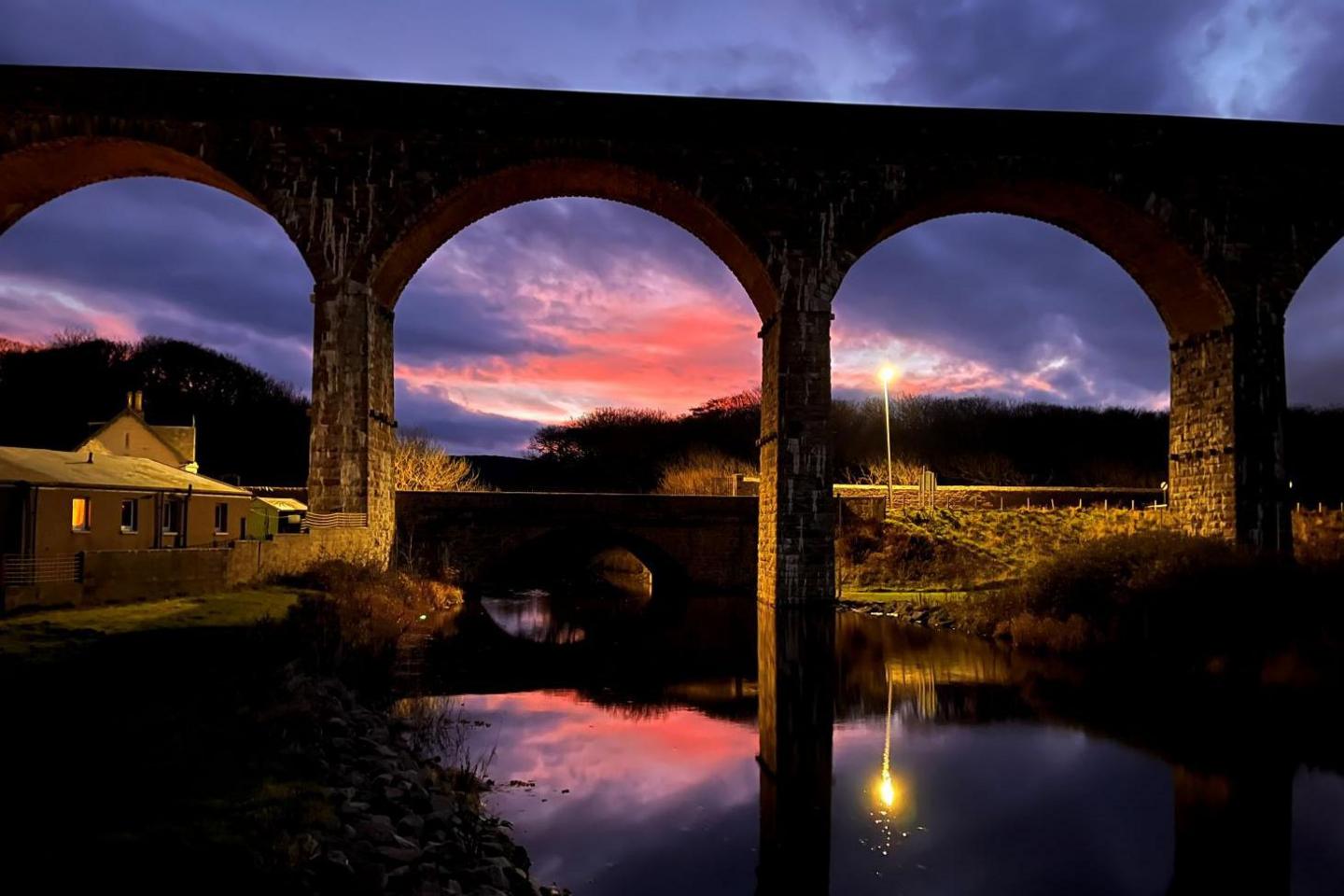 The viaduct is reflected in water below. There is a house with lights on nearby and there are clouds of dark blue and orange in the evening sky.