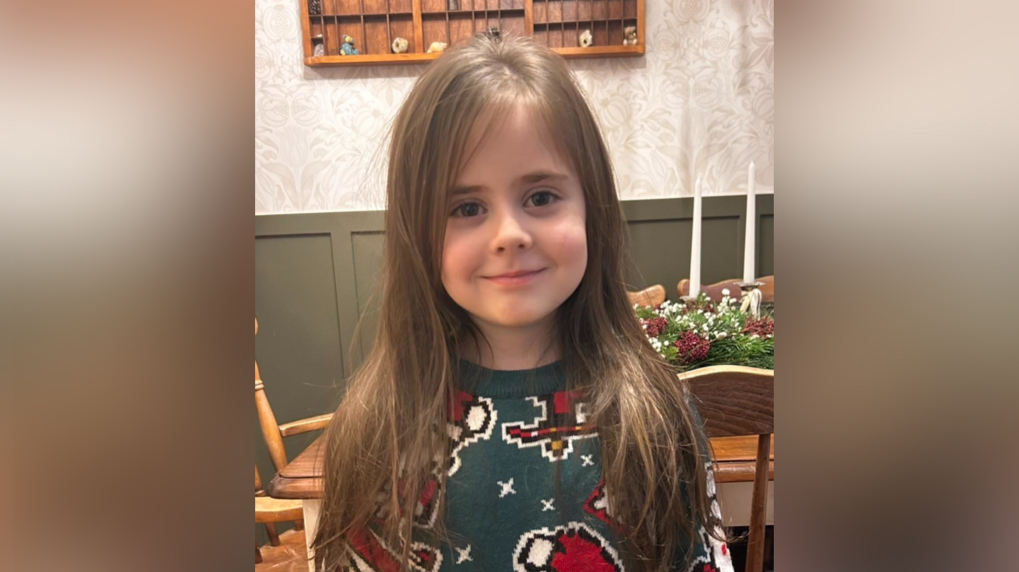 Reuben smiles at the camera. He has long brown hair that is loose. He is wearing a green jumper with Christmas cartoon characters on it.