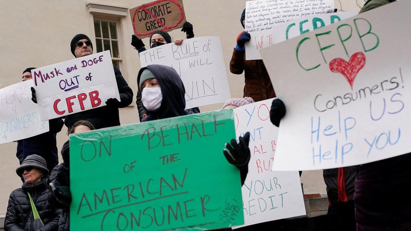 A group of protesters holding signs with slogans including "Musk and Doge out of CFPB" and "On behalf of the American consumer" 