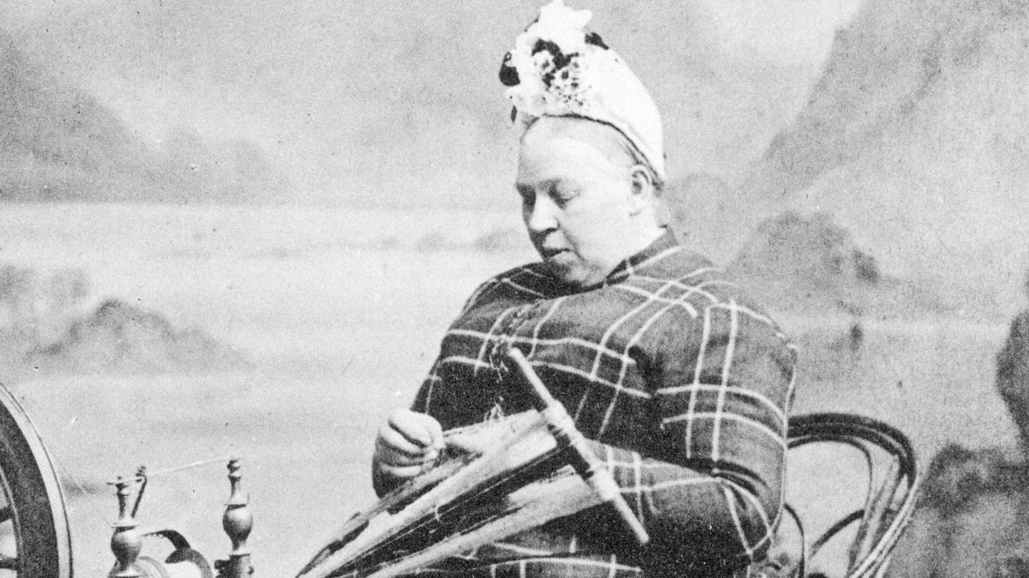 A woman sits at a spinning wheel weaving thread. She is dressed in tartan and wearing a hat.