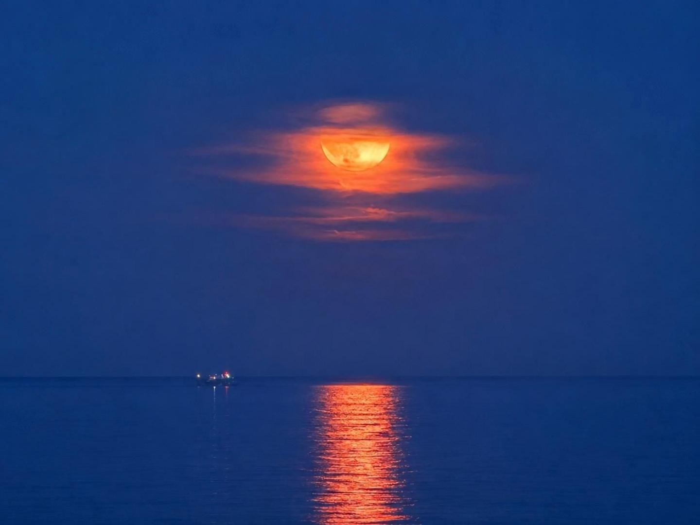 An orange hued full Strawberry Moon appears in a blue night sky above a still blue sea with a boat floating in it. The sea reflects the moonlight.