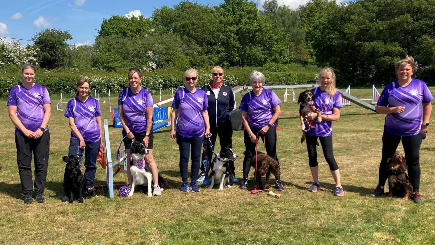 The seven competitors are pictured wearing their purple club tops. Six of the women have their small and medium sized dogs with them. They are stood in an agility field with the apparatus visible behind them.