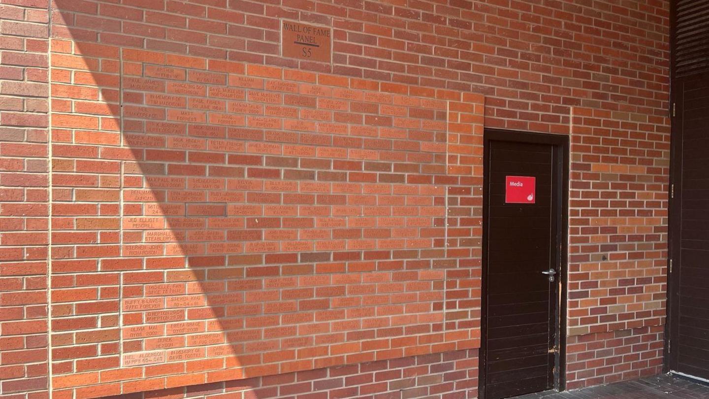 A wall of brown bricks. More than 100 of the bricks have names written on them. A larger block at the top of the section has Wall of Fame Panel S5 written on it. There is a black door next to the panel, with a red media sign on it.