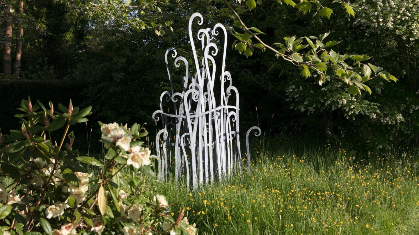 A grey metal sculpture depicts grass fronds emerging from the ground in front of green shrubs and trees