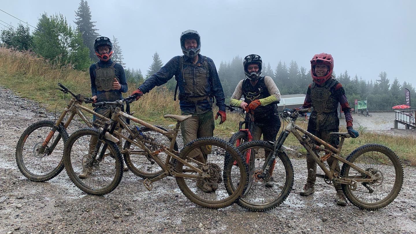 A group photo of the family at a wet trail centre, stood with their bikes. They are covered in mud head to toe. They're wearing full-face helmets but you can tell they're all smiling.