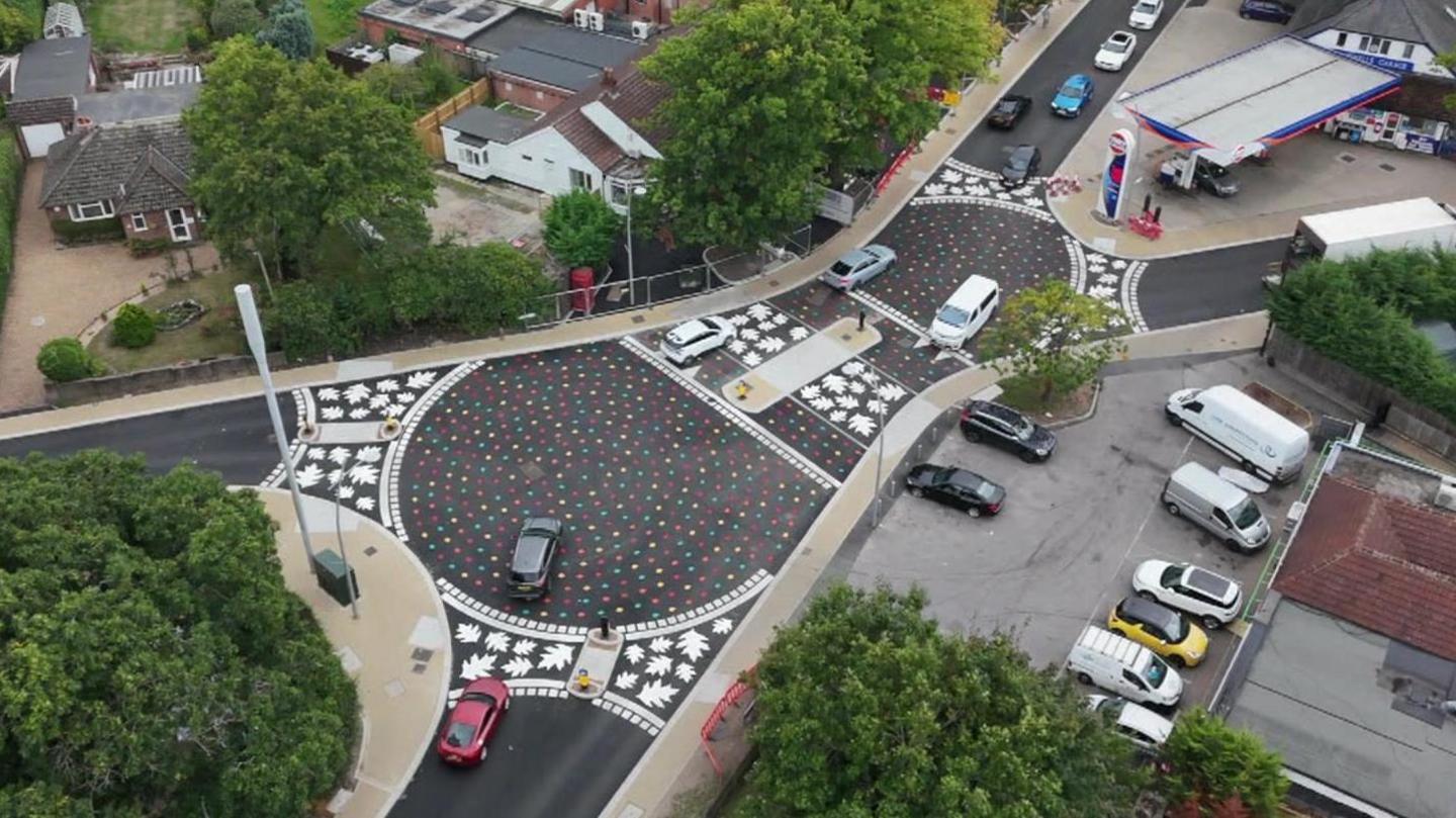 An aerial photo of two roundabouts, with rainbow leaves painted on the tarmac. At each of the the three exists for each roundabout there is a band of white leaves painted across the road.