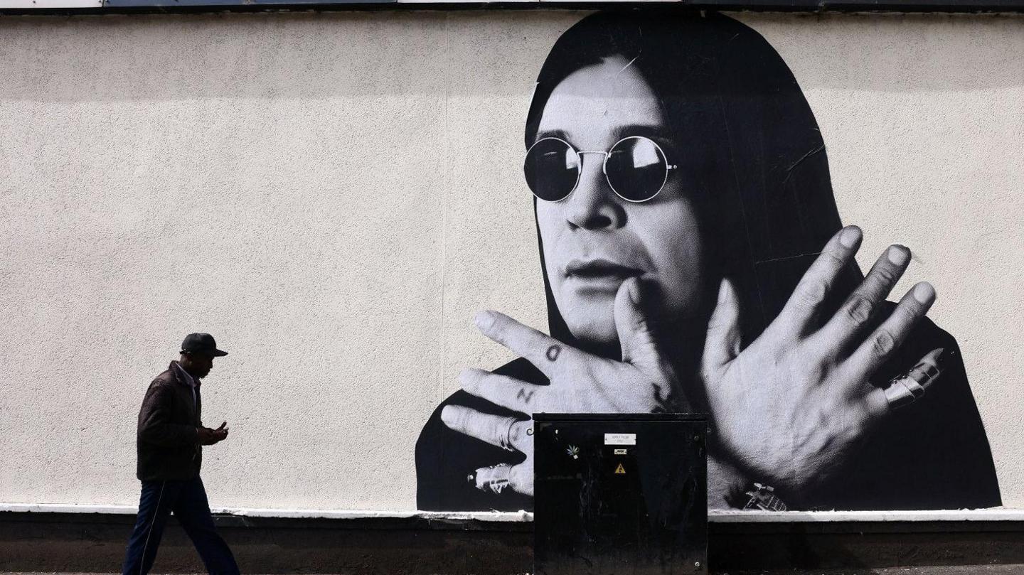 A large wall mural of Ozzy Osbourne, who has long dark hair and dark circular sunglasses. His hands are crossed in front of him, his fingers are splayed out. He has knuckle tattoos on one hand, reading "Ozzy".