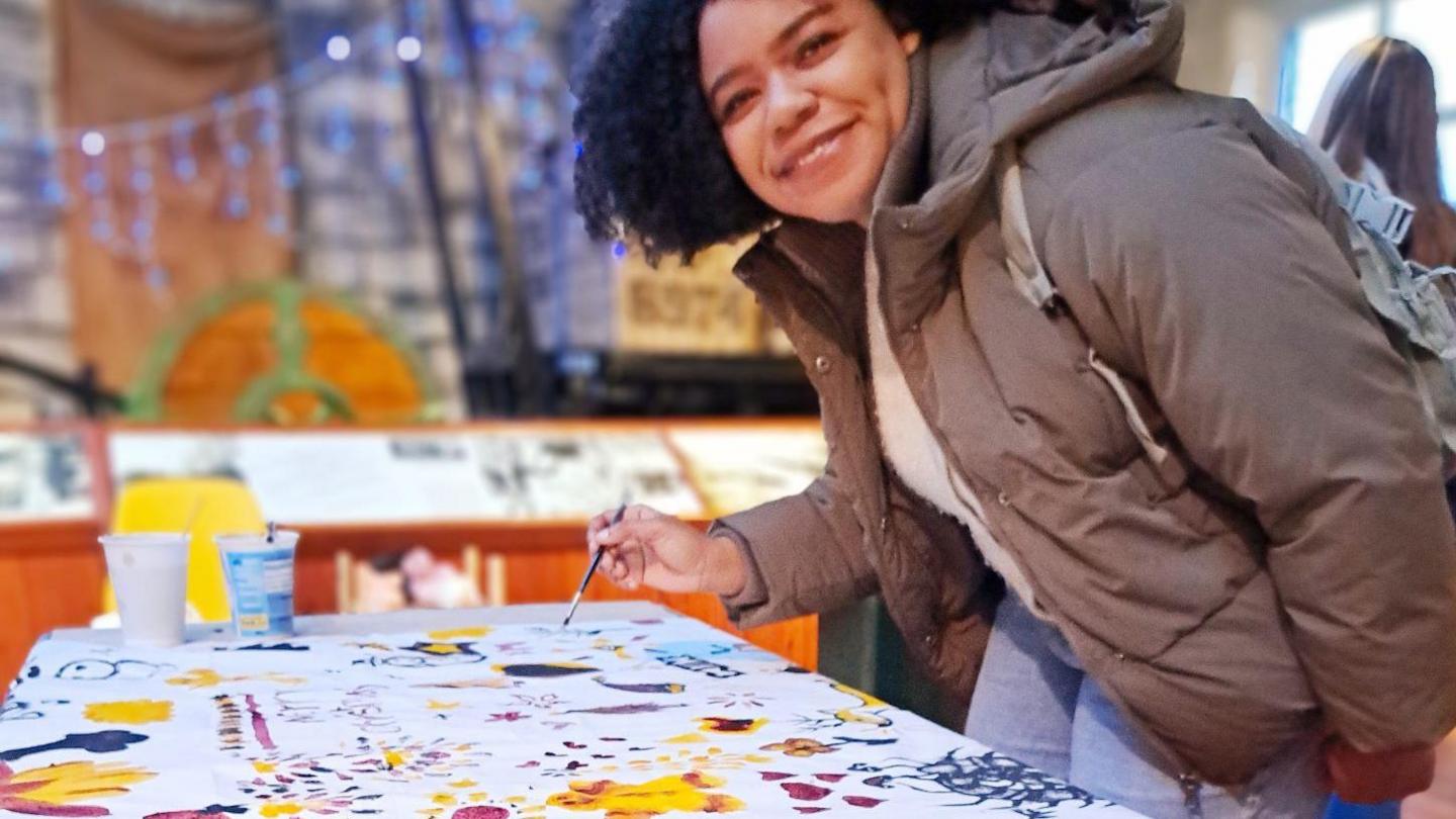 A woman with dark hair and waring a grey coat, jumper, jeans and a backpack is holding a paintbrush. She is leaning over a cloth on a table. The cloth is decorated with patterns and pictures in bright colours.