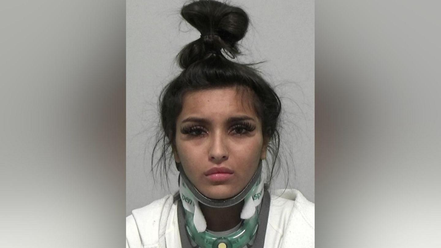 Mugshot of a woman wearing a neck brace, with remnants of make up on her face and her long black hair tied in a knot atop her head