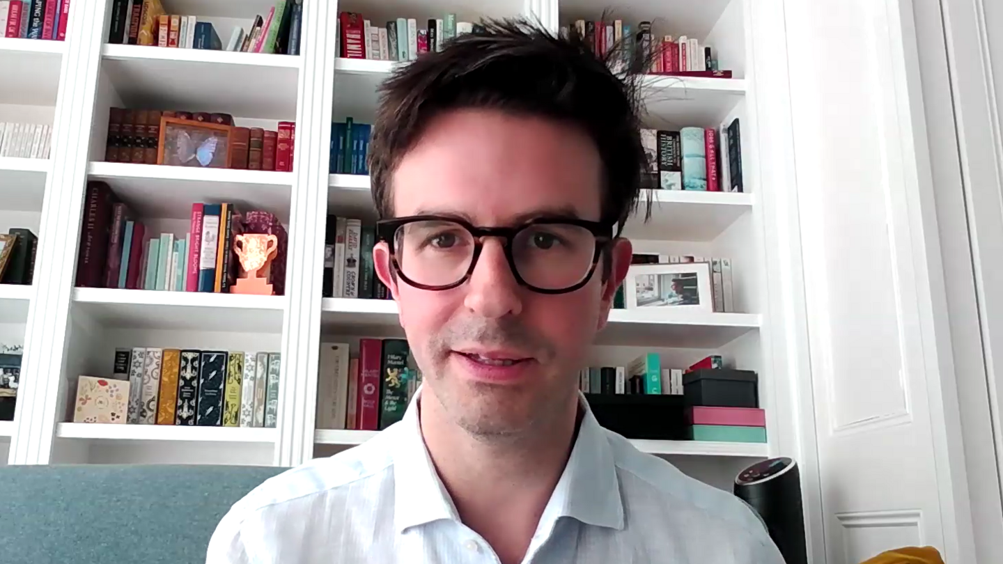 A man, Aron Gelbard, is seated on a sofa in front of a bookshelf filled with various books and decorative items. He is wearing a light-colored shirt, and a yellow pillow is visible to his right. The background is in focus, showcasing the bookshelf’s contents clearly.