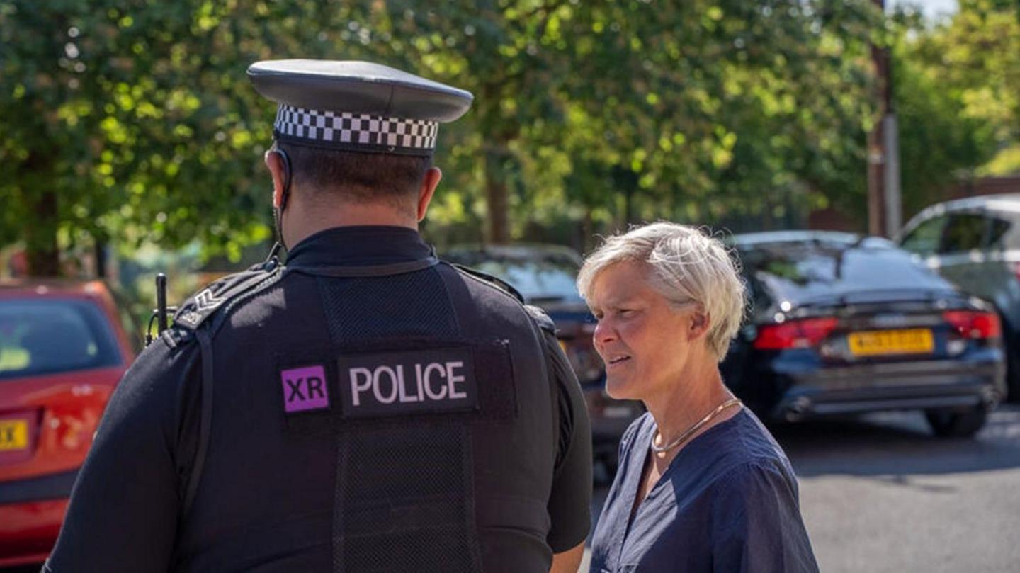 A woman with short blonde hair appears to be in conversation with a police officer in a car park 