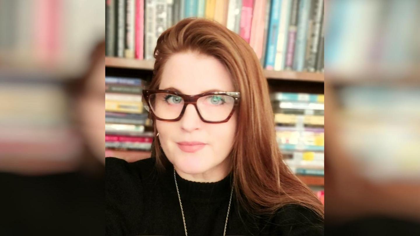 A woman with strawberry blonde hair and big, brown glasses. Behind her is a full bookcase. 