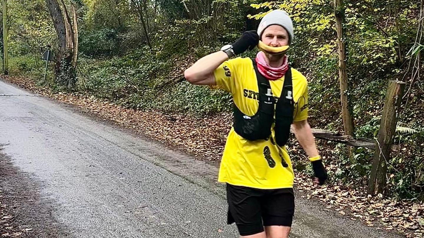 a man on a road in running gear and a beanie hat holds a banana to his mouth as a smile