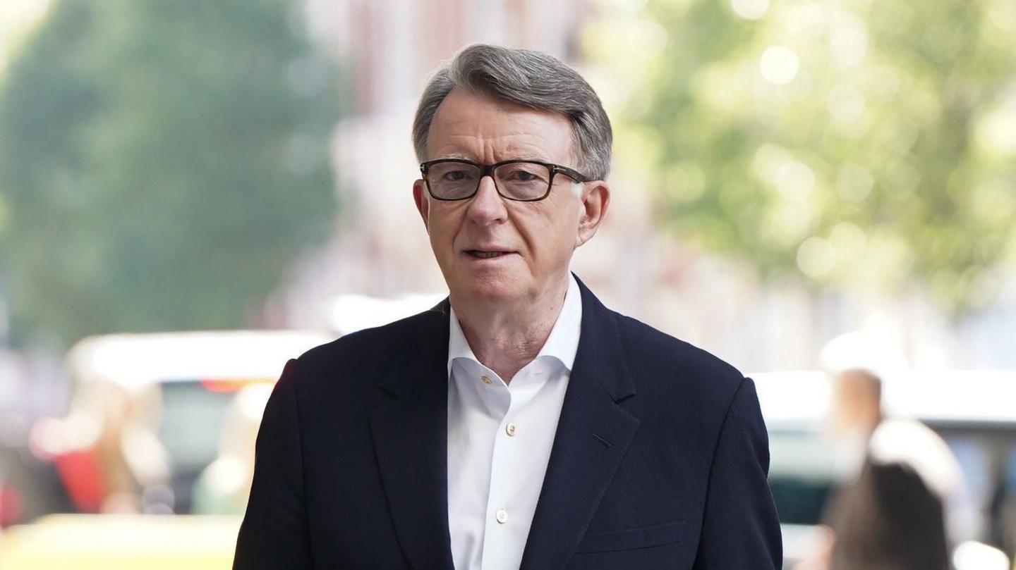 Lord Mandelson. A grey-haired man with black glasses is walking, wearing a white shirt and navy suit jacket. He has a neutral expression on his face.