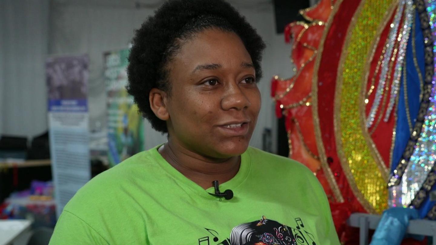 A woman with short curly black hair wears a green T-shirt. She is looking away from the camera. Behind her is a colourful, sequined outfit.