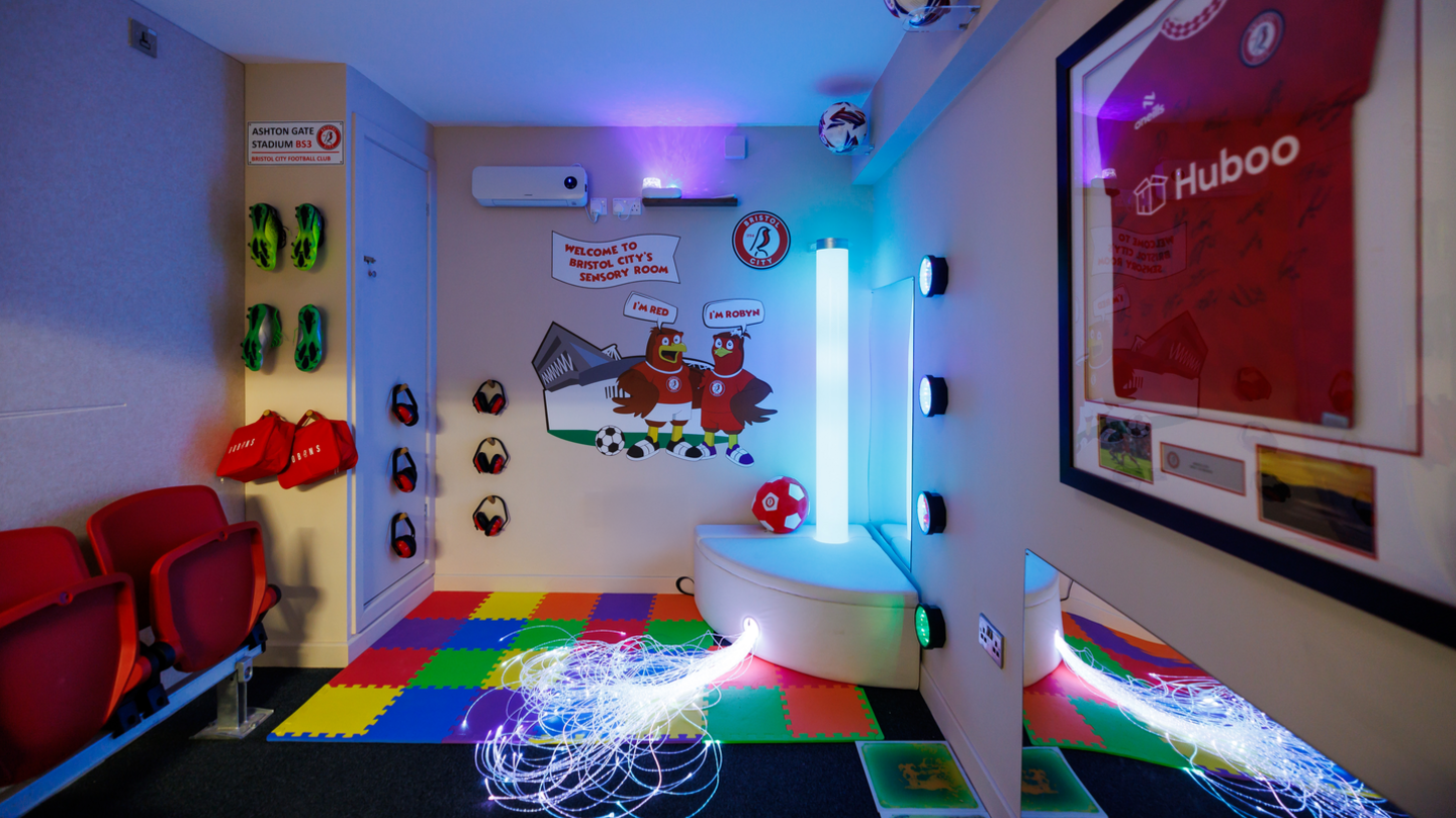 A sensory room featuring - from left to right - plastic seating, football boots hung on the wall, headphones, a mural of two robins, a light display and a signed Bristol City shirt.