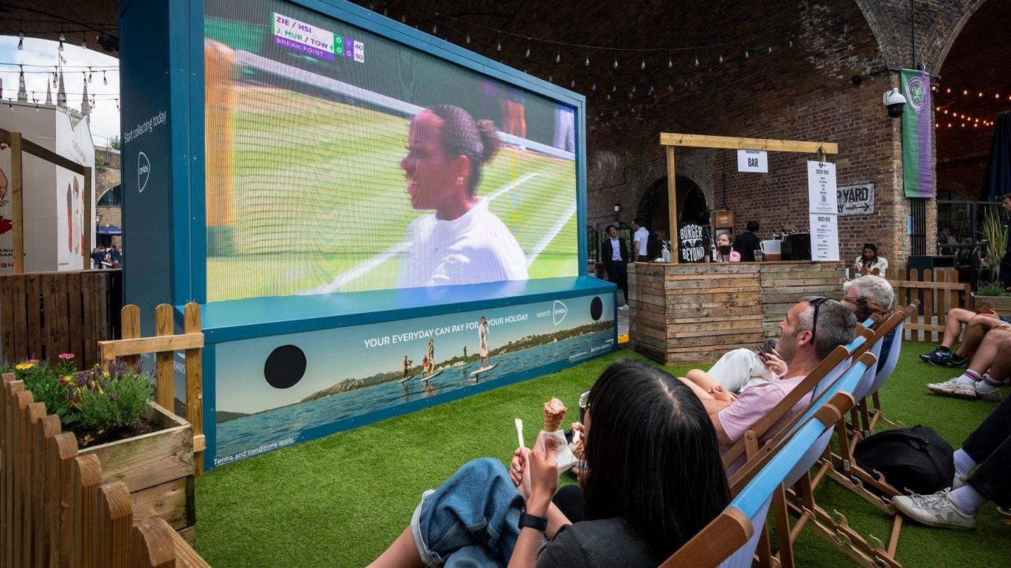 A photograph of a big screen being used outside to show a tennis match, with people watching in deckchairs. There is a bar to one side of the screen and people are wearing sunglasses and eating ice cream.