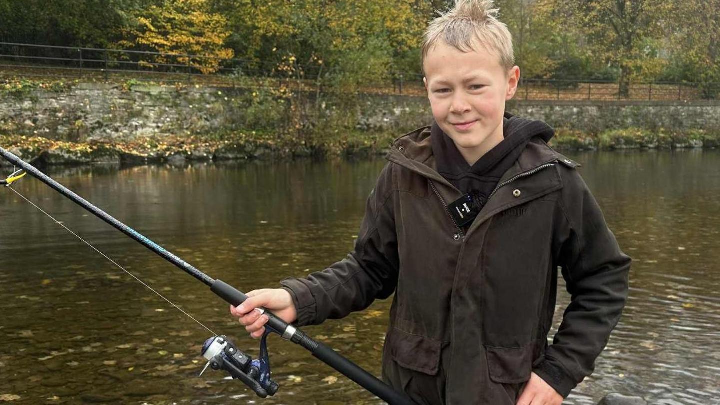 Bailey, a young boy with blonde hair, wears a brown wax coat as he casts a fishing line from his rod into the River Kent. It is a grey rainy day and the visibility is poor.