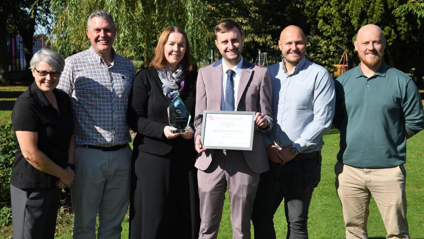 Six members of the wellbeing team standing in line for a photo, with a young man wearing a suit, shirt and tie holding the award