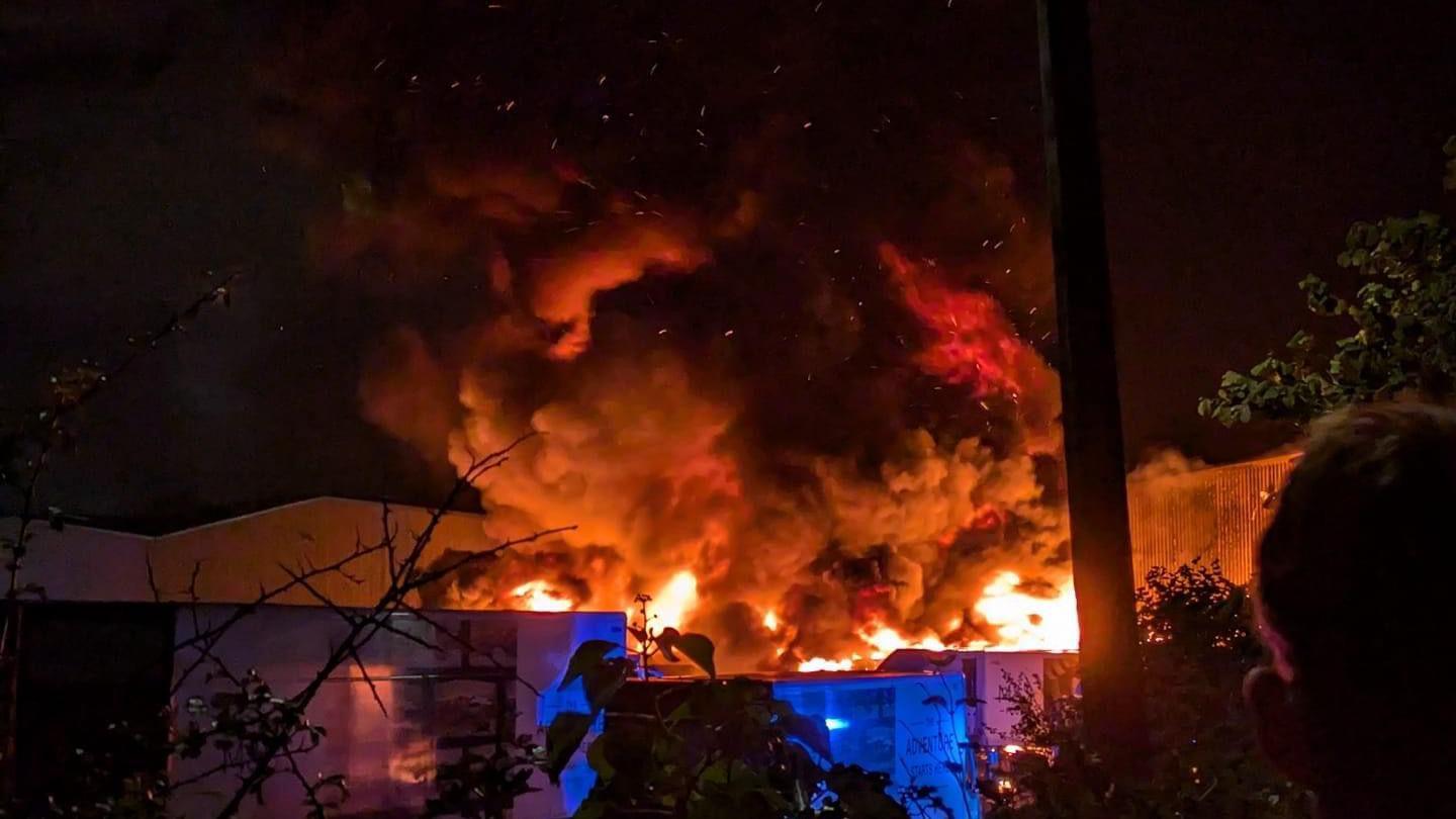 A huge fire at an industrial estate. Orange flames and a thick plume of smoke can be seen. 