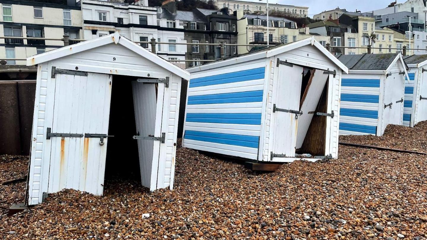 A line of beach huts all scattered across a stony beach and not where they are supposed to be. They are blue and white. The pier can be seen in the back ground. 