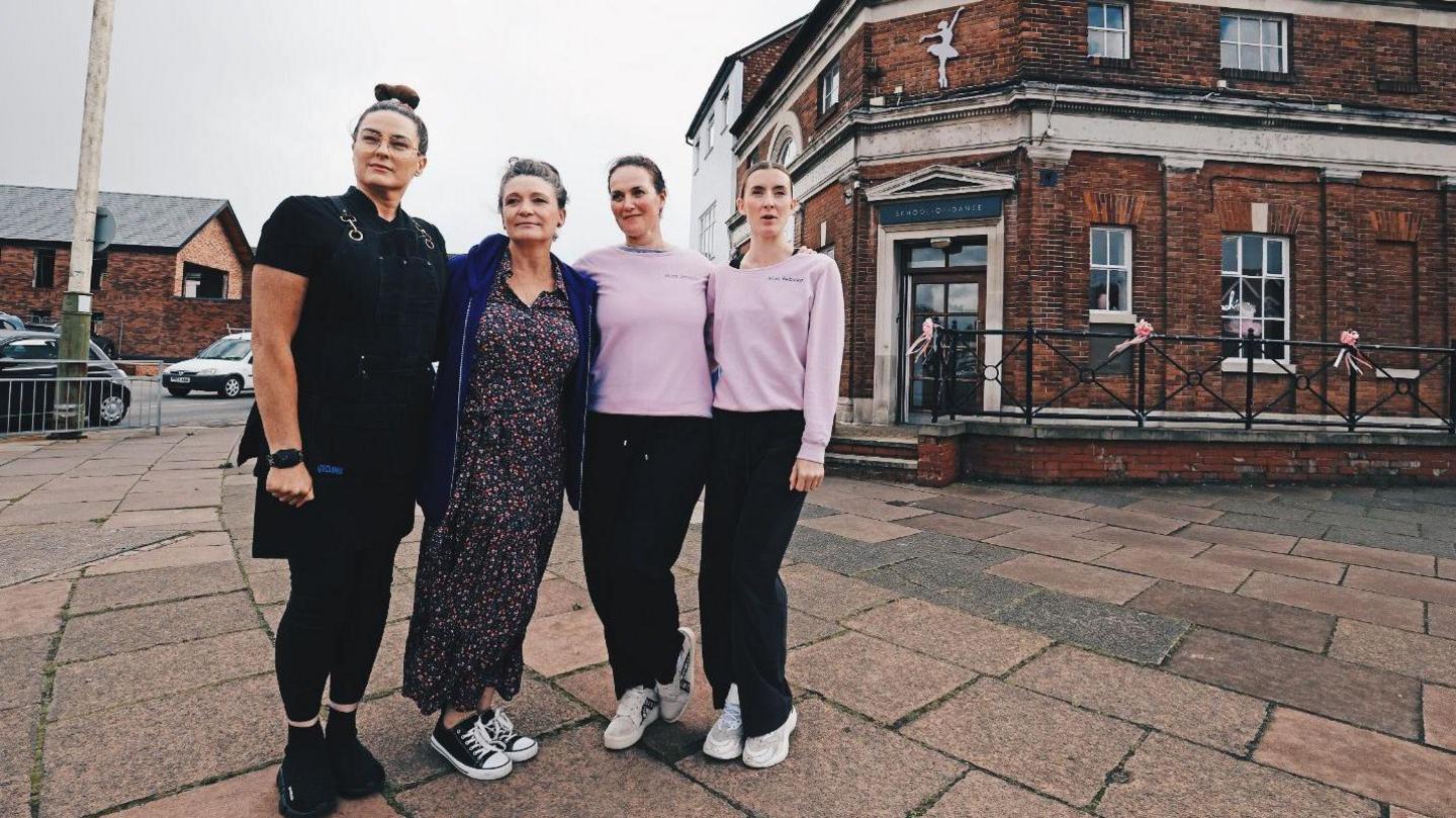 A woman wearing a black top and skirt, stands next to another woman wearing a floral dress, and two other women both wearing identical pink tops and dark trousers. They are in front of a building which has a ballerina image on the front and a sign over the door that says 'School of Dance'.