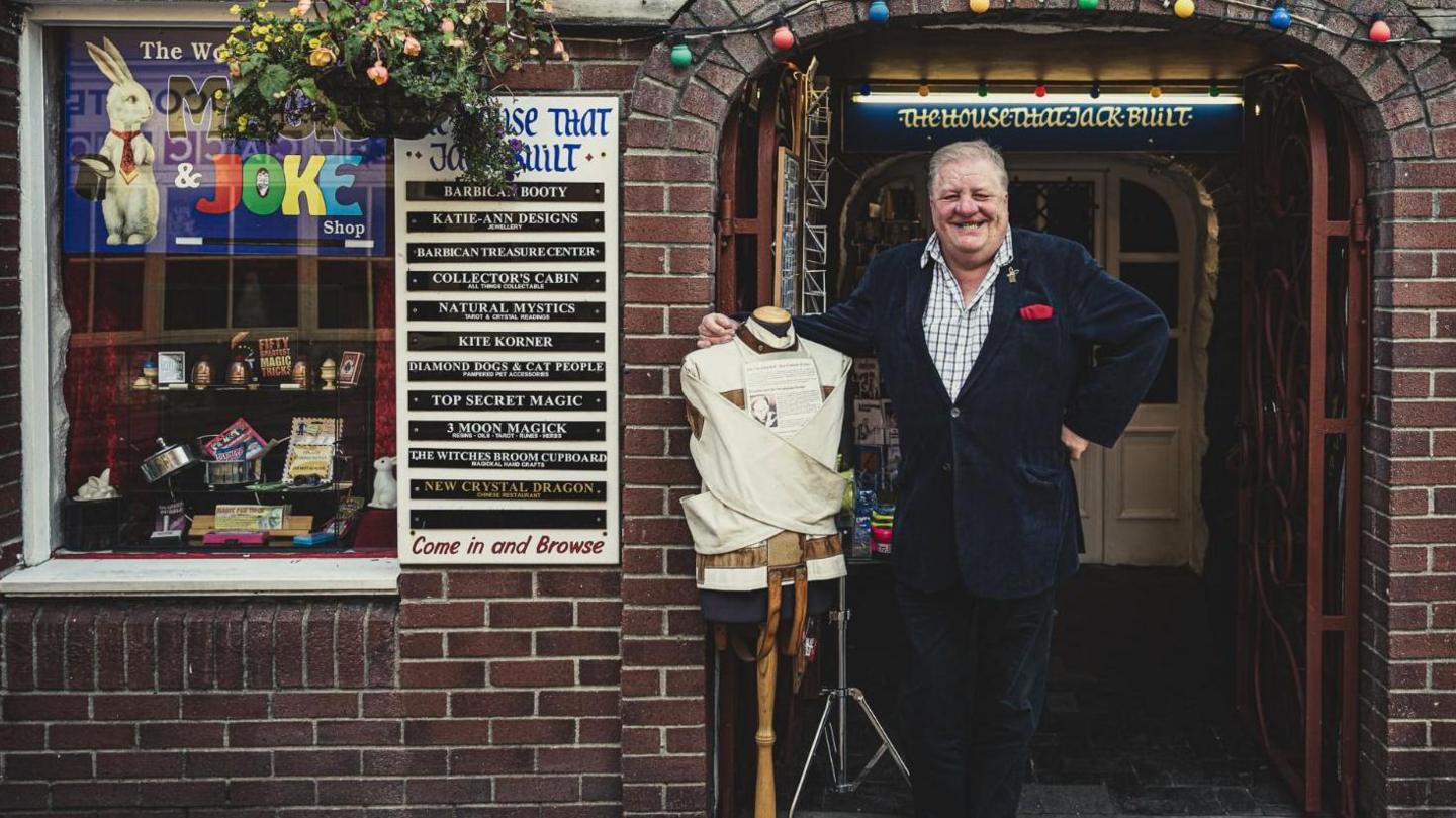 Older photograph of Malcolm Norton stood outside his shop with the straitjacket on a mannequin. Mr Norton is clean shaven and smiling at the camera with his right arm resting on the mannequin. He is wearing a dark blue jacket and dark coloured trousers.