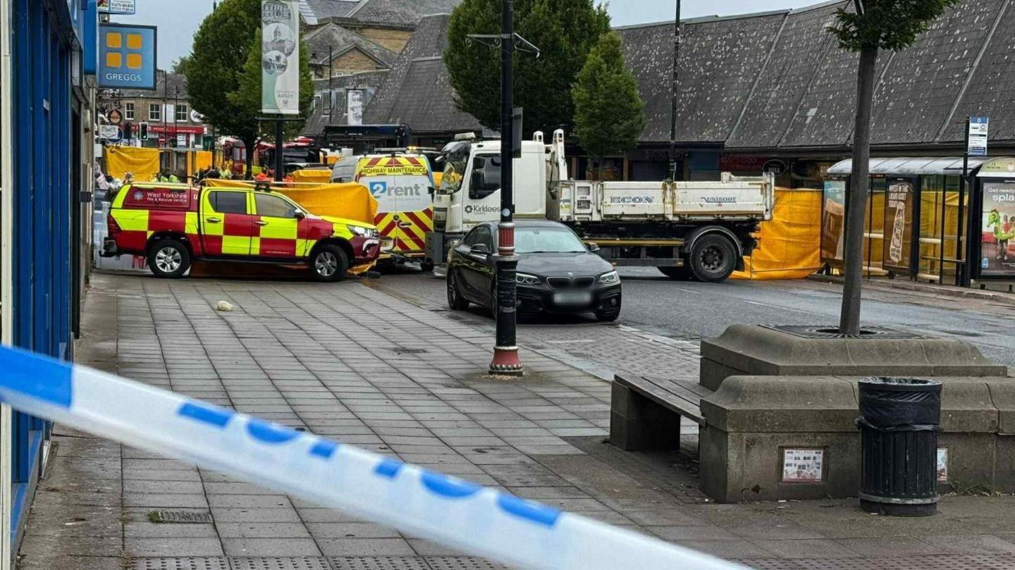 A street which has been cordoned off with police tape and there are several vehicles within the cordon, including trucks and a fire service vehicle.
