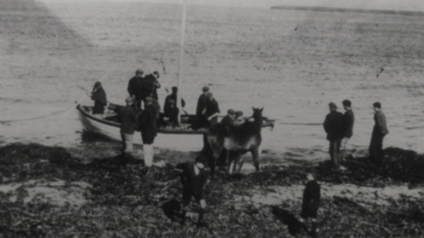 A black-and-white photograph of a boat at the seashore with men inside her and a horse with other men waiting to board.