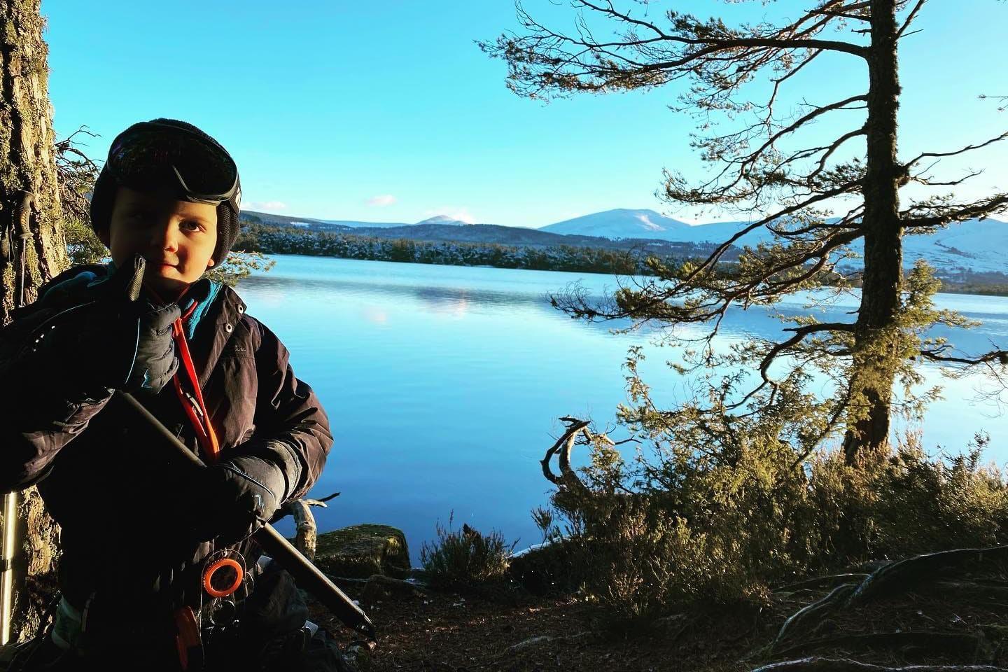 Rio by a lake with some scenic mountains in the background. He is wearing many layers of outdoor clothing and is holding his thumb up to the camera.