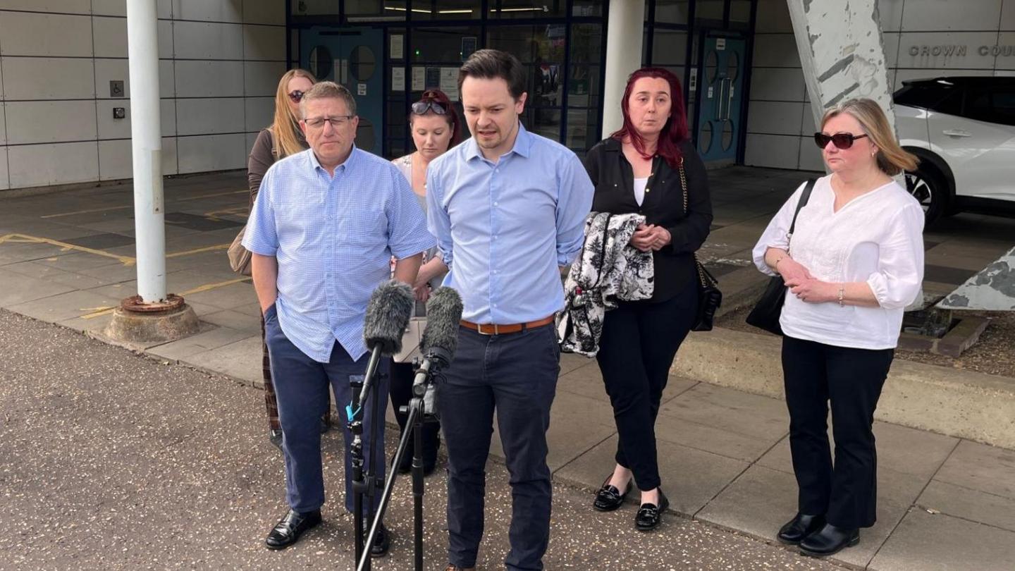 A group of six people outside a courthouse. One man is speaking into two microphones on stands.