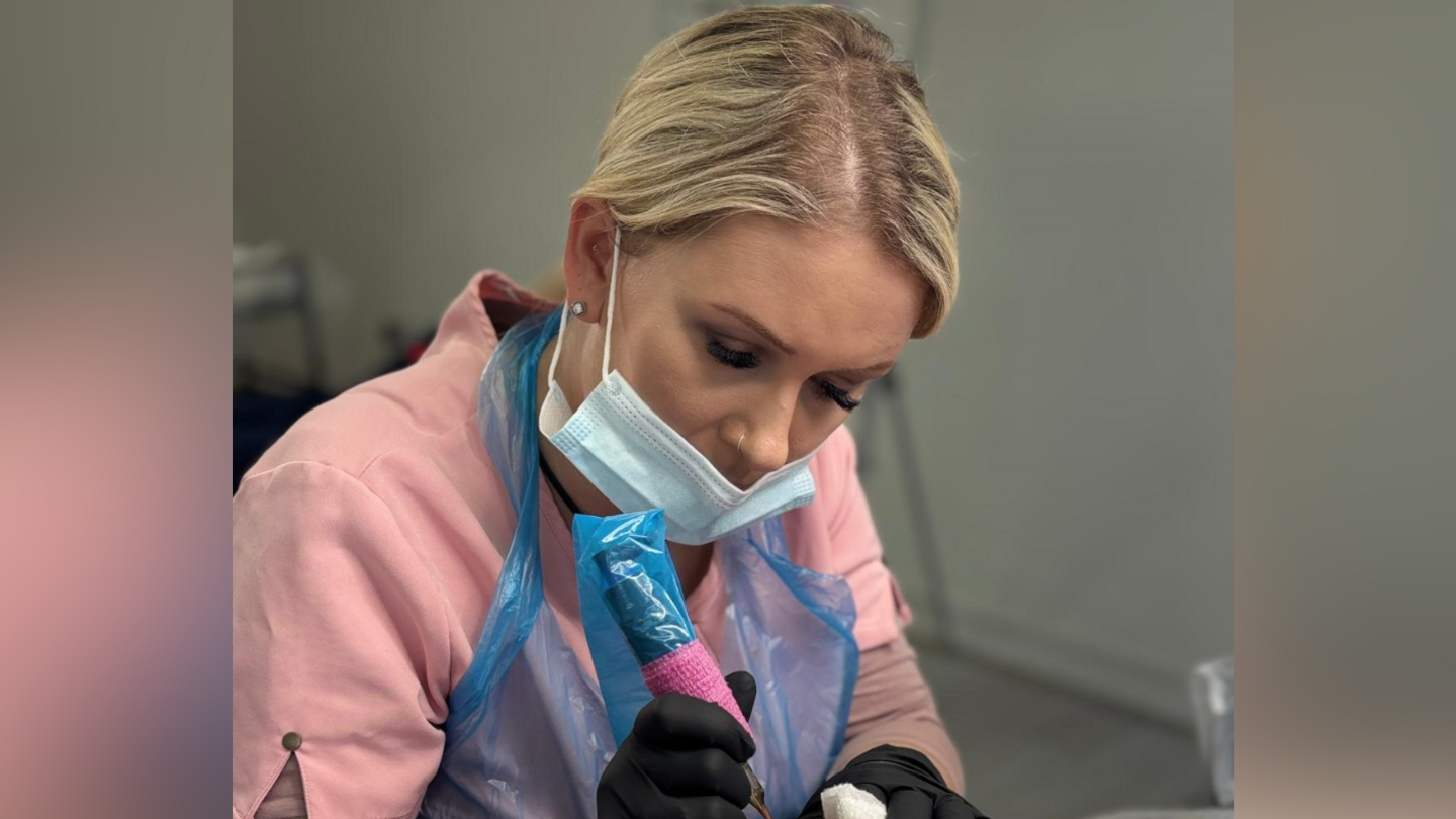 Kirsty Jackson is pictured using a tattoo gun. She wears a blue mask on her face, black gloves and pink uniform with a blue plastic overall over the top. Her blonde hair has been tied back.