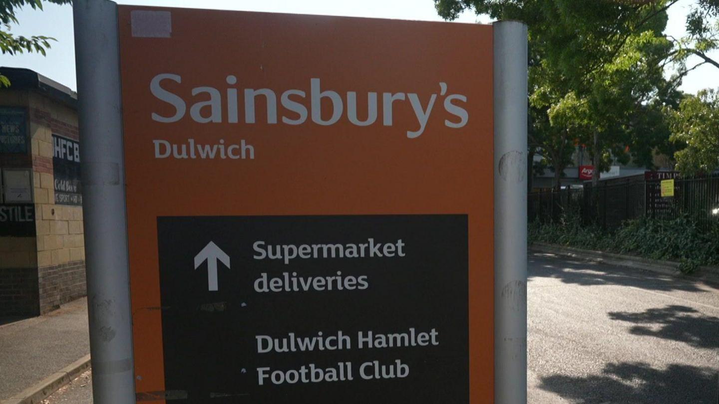 An orange and black road sign reading "Sainsbury's Dulwich" with an arrow pointing straight ahead for "Supermarket deliveries" and "Dulwich Hamlet Football Club".