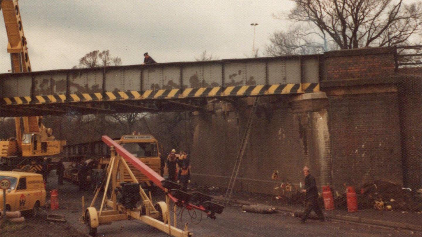 The colour photograph from 1975 shows the scene at the A5 bridge in Hinckley. Engineers are there and a crane and other vehicles and equipment can be seen. They are making a temporary repair. A year later a new bridge section was installed. 