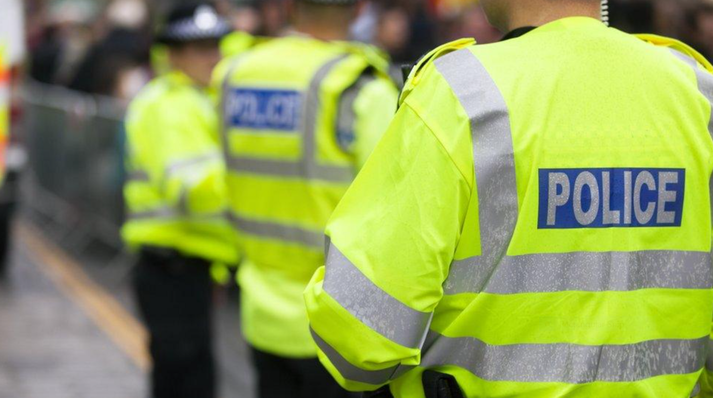 A police officer wearing a high-vis jacket facing away from the camera. Other police officers can be seen in the background but they are blurred. 