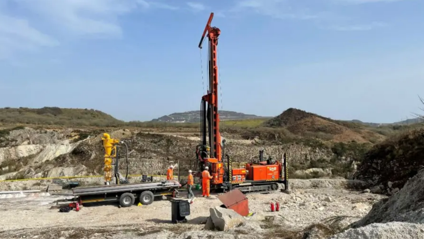 A drilling rig clad in orange coloured external metalwork sits atop a landscape which appears to show a history of mineral extraction evidenced by the hinterland of large pits and piles of what appear to be waste material, alongside three people attired in orange clothing and wearing hard hats.