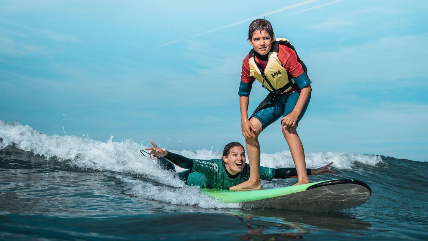 Vicky lies on a surf board as she rides a small wave with a child standing on the board