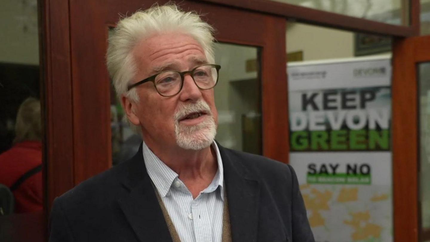 A wide shot of Stephen Crowther. He has short grey hair, is wearing glasses, a white shirt, and a dark jacket.