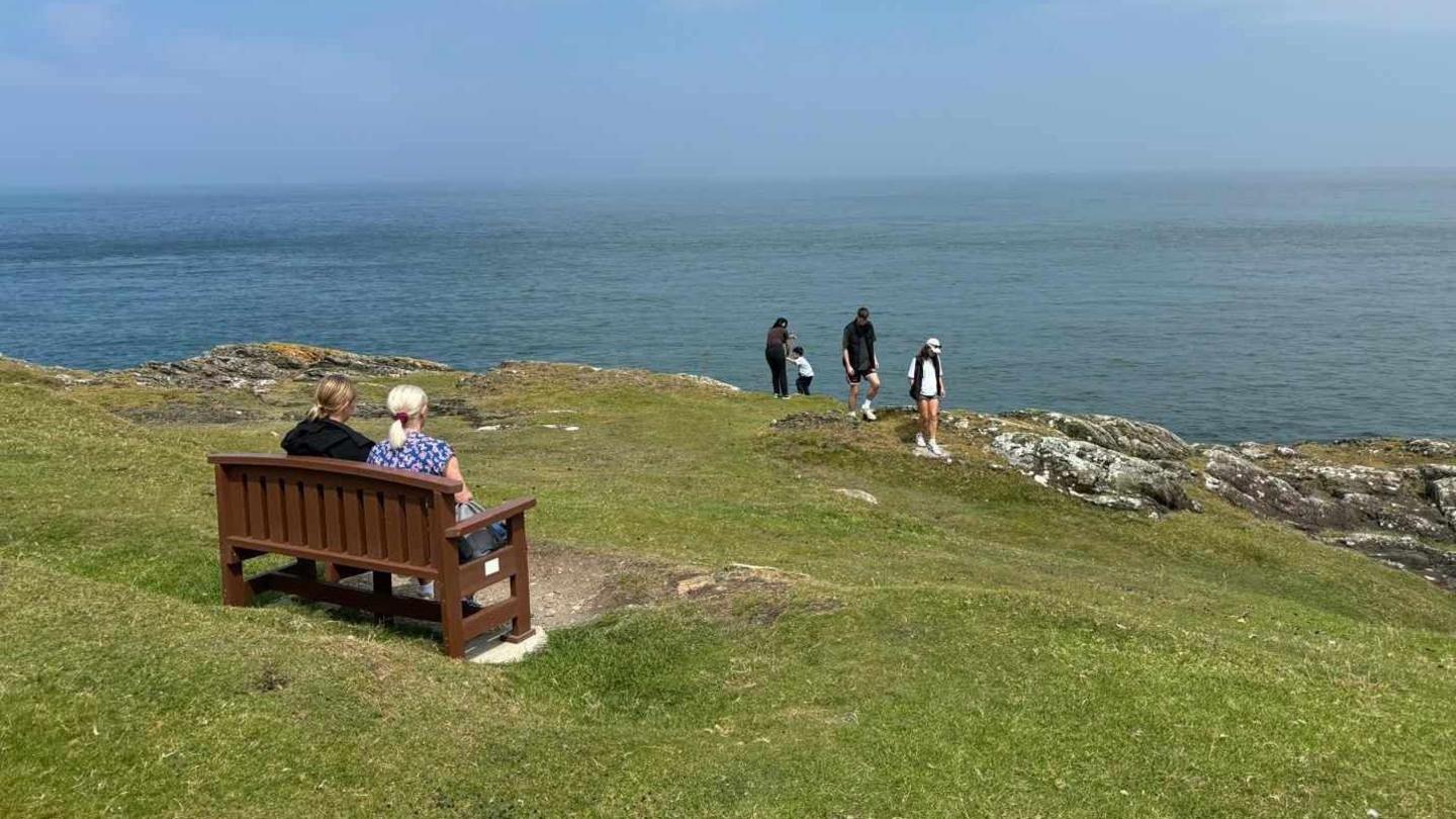 Two women sit on a brown wooden bench placed on a concrete base on a green headland looking out to sea. A family can be seen walking on rocks at the bottom of the area. 